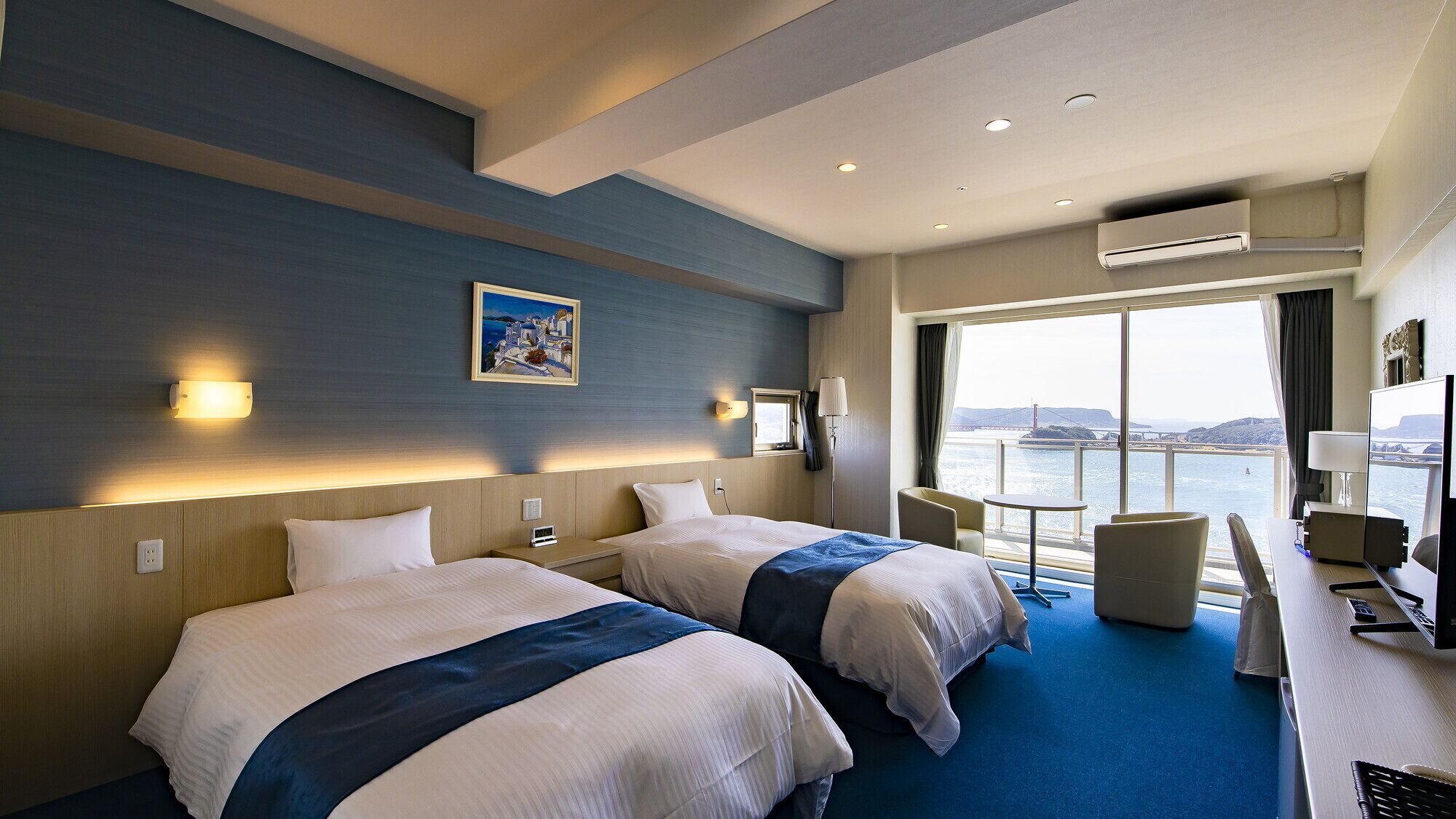 Habitación de hotel con dos camas, un escritorio con silla, una mesita y un amplio ventanal con vistas a un puente y al agua.