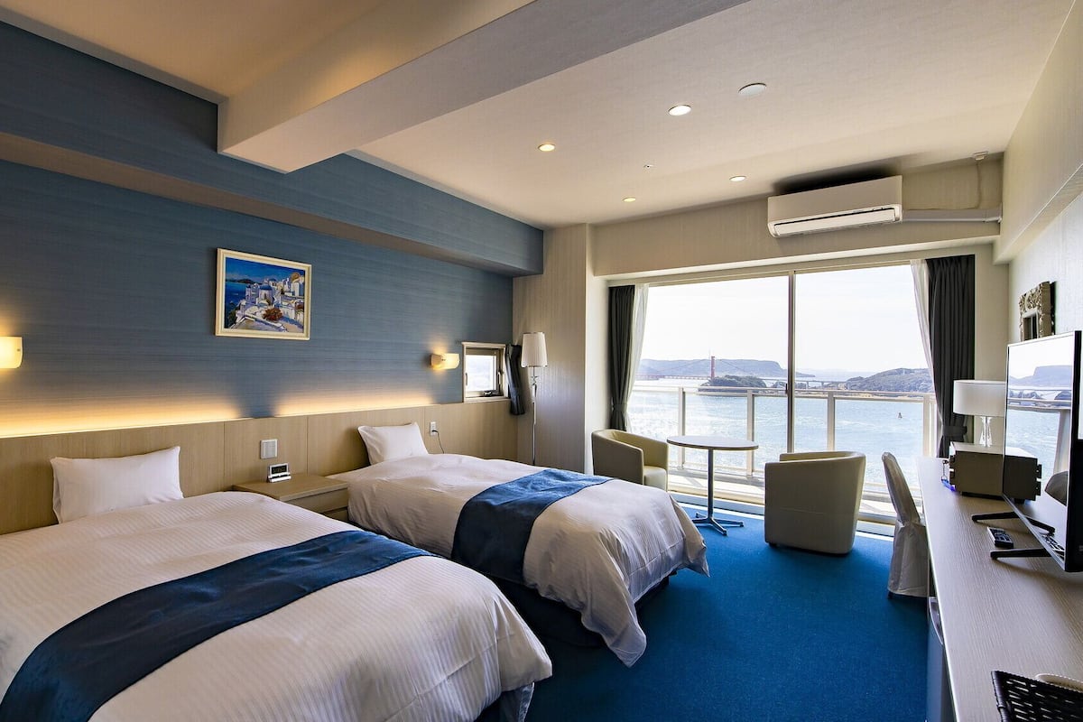 Habitación de hotel con dos camas, un escritorio con silla, una mesita y un amplio ventanal con vistas a un puente y al agua.