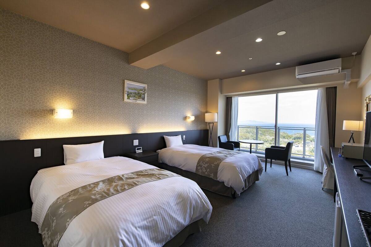 Habitación de hotel con dos camas, un escritorio, una silla y un ventanal con vistas.
