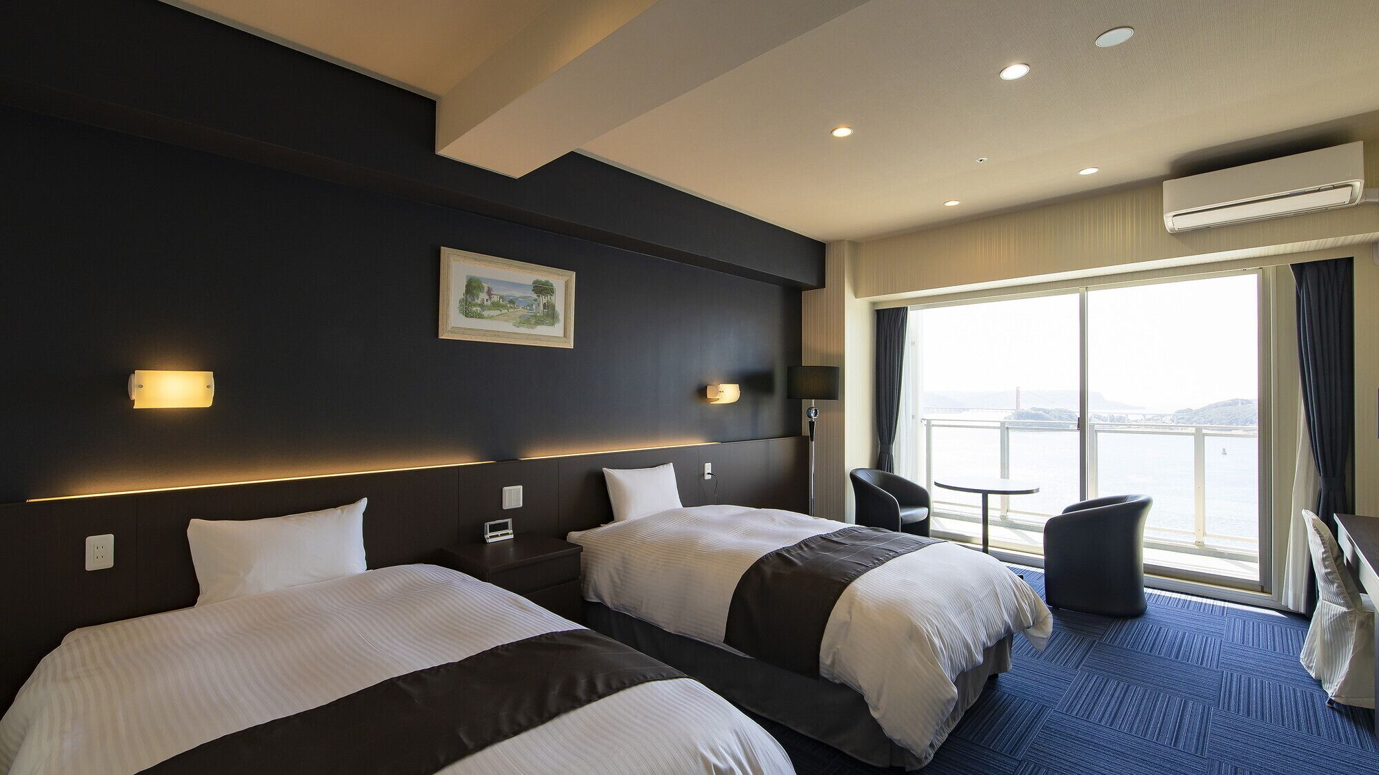 Habitación de hotel con dos camas, un escritorio, una silla, un ventanal con vistas y un equipo de aire acondicionado.