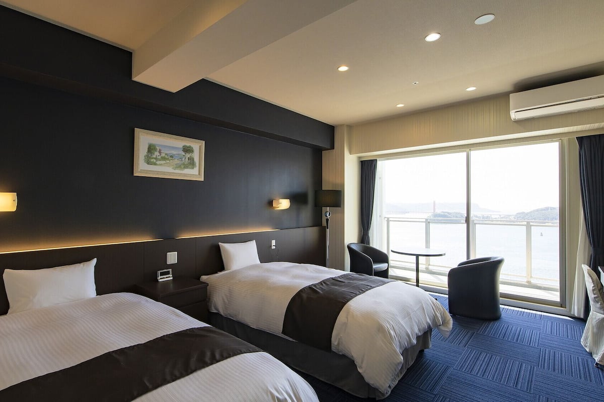 Habitación de hotel con dos camas, un escritorio, una silla, un ventanal con vistas y un equipo de aire acondicionado.