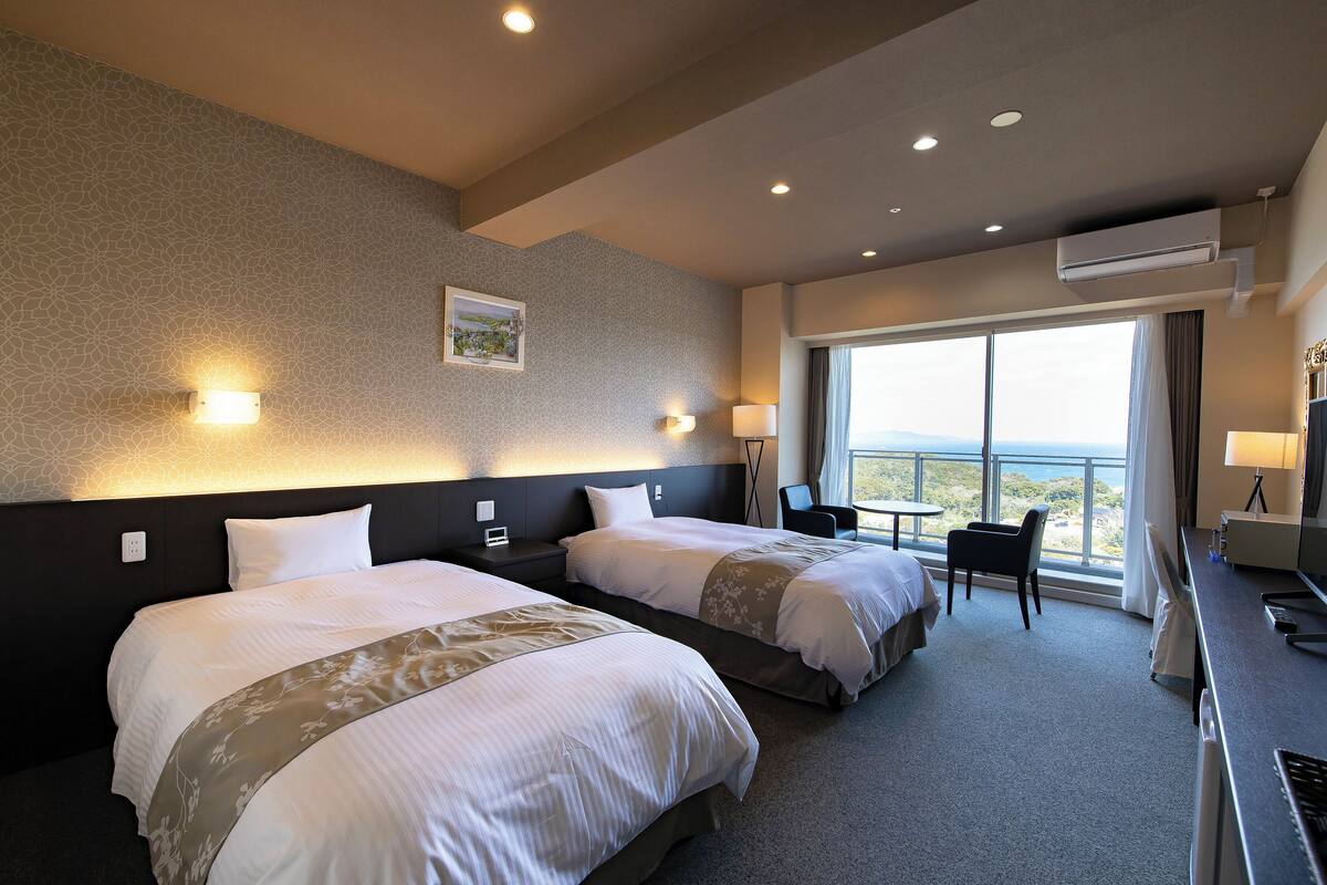 Habitación de hotel con dos camas, un escritorio, una silla y un ventanal con vistas.