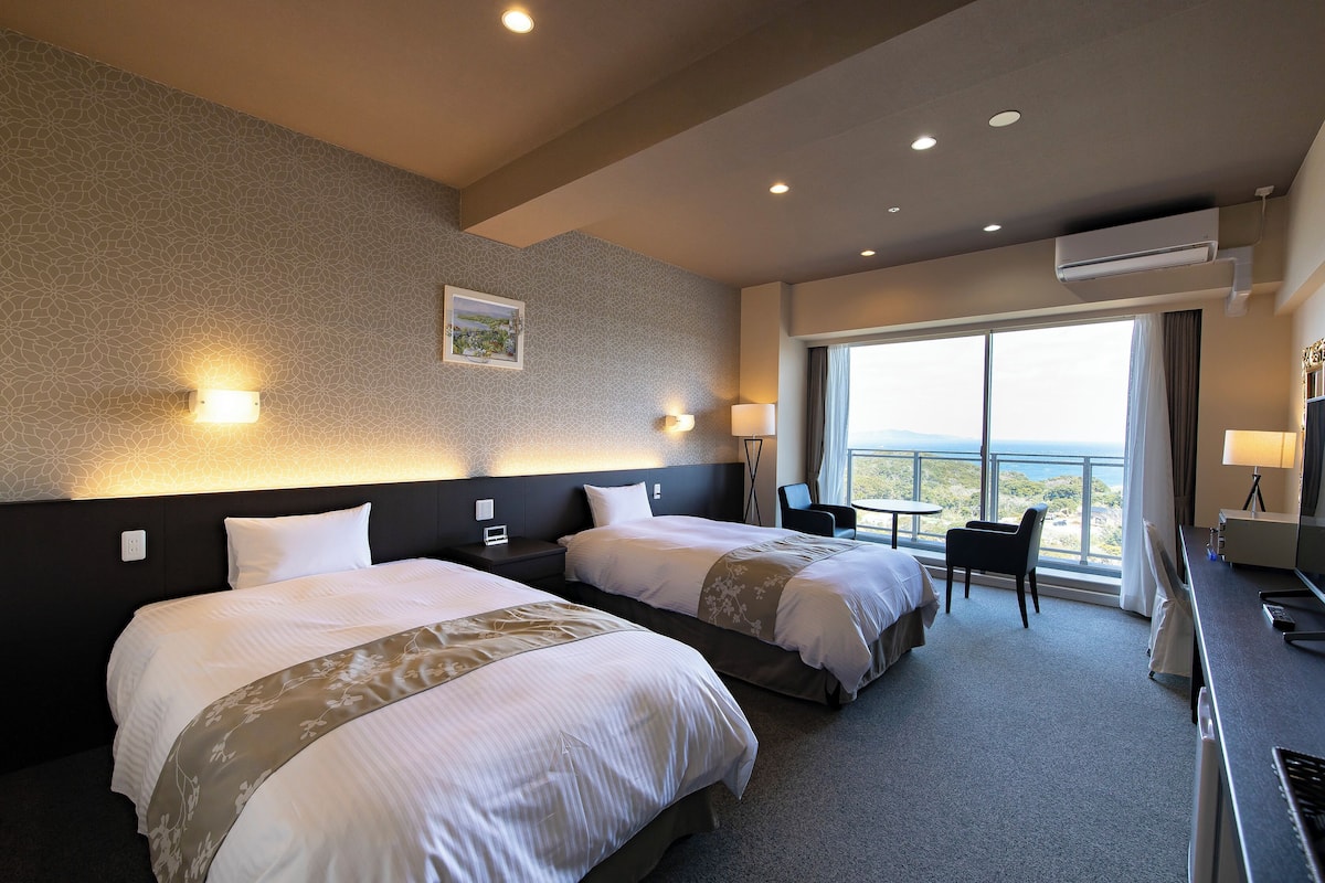 Habitación de hotel con dos camas, un escritorio, una silla y un ventanal con vistas.