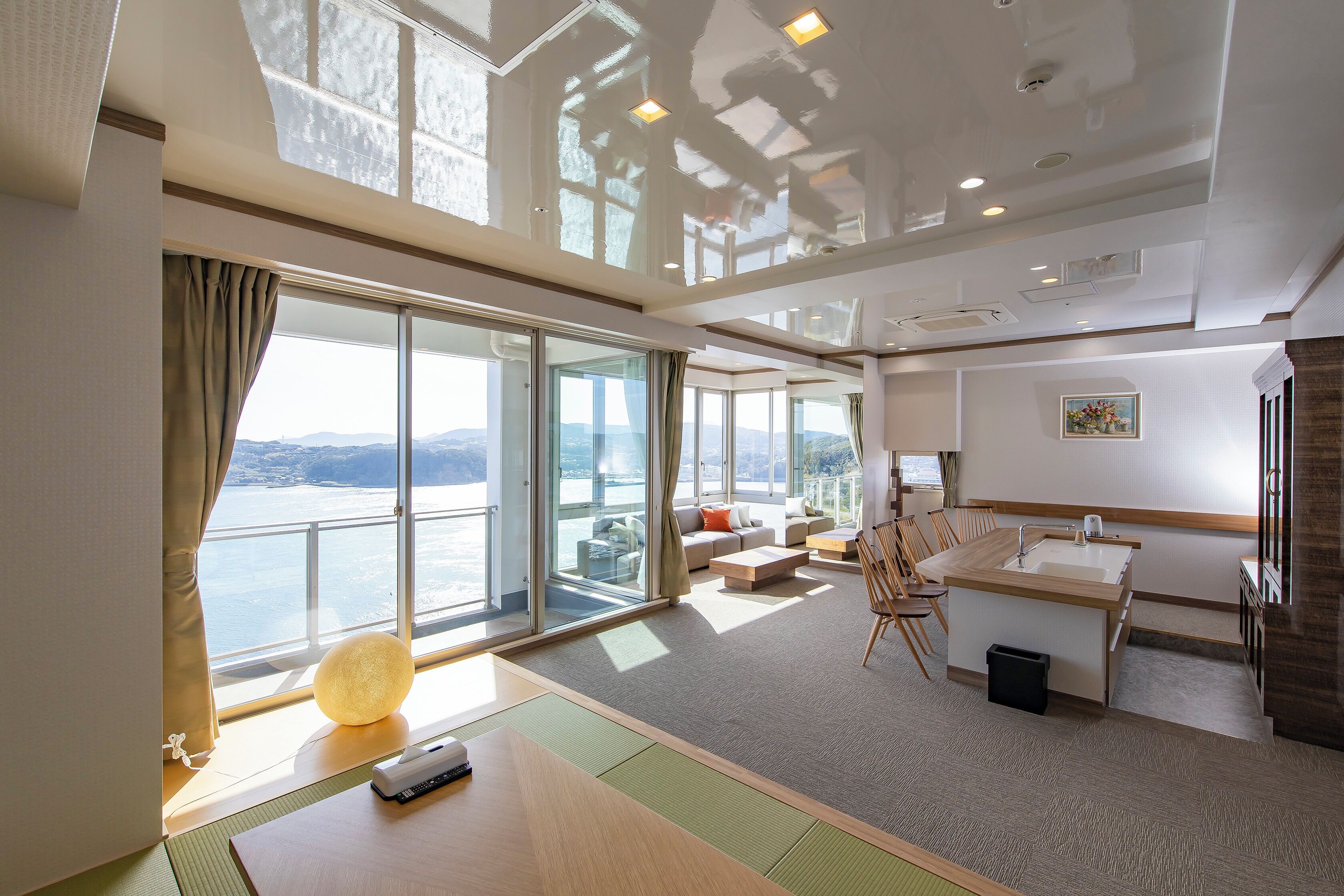 Una habitación de hotel moderna con un gran ventanal corredero a un balcón, un área de descanso con sofá y sillones, una pequeña cocina y vistas al mar.