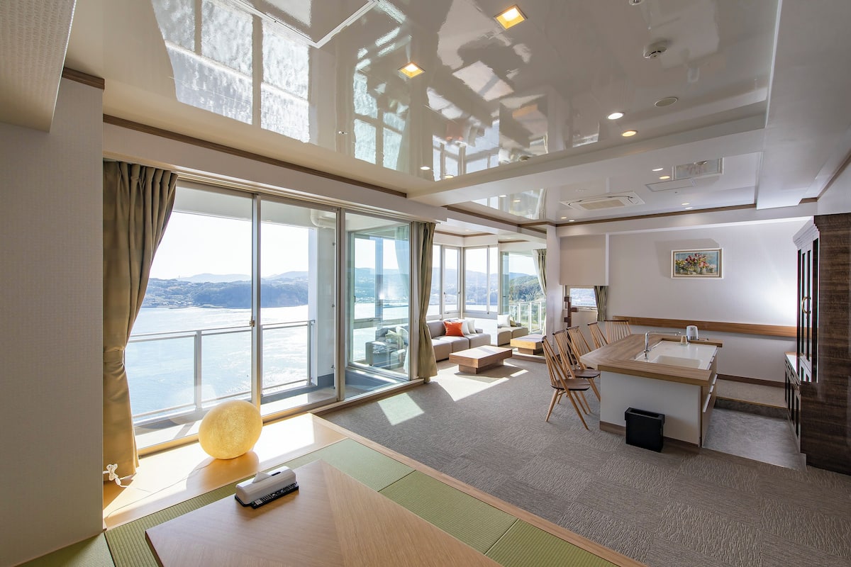Una habitación de hotel moderna con un gran ventanal corredero a un balcón, un área de descanso con sofá y sillones, una pequeña cocina y vistas al mar.