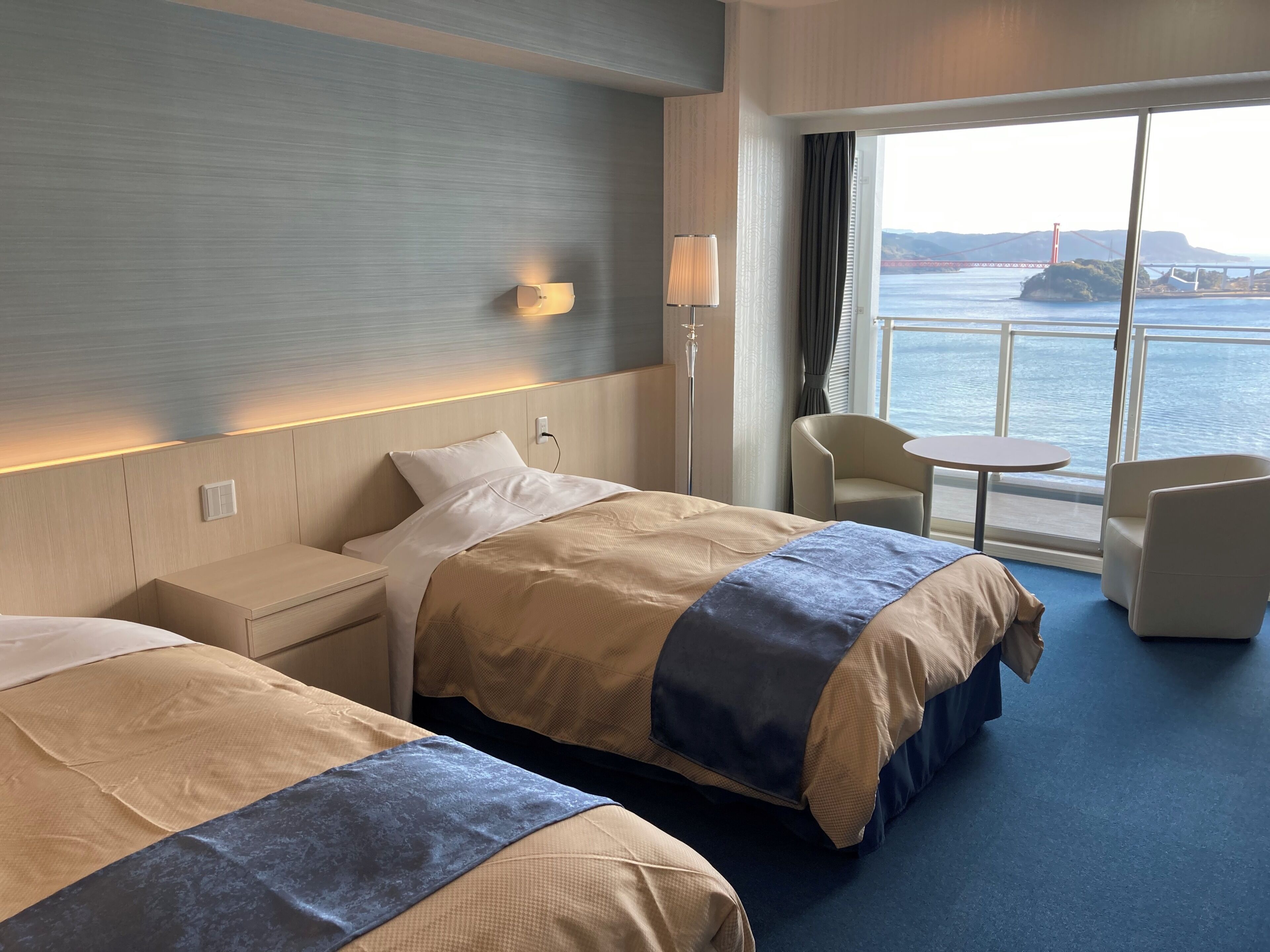 Habitación de hotel con dos camas, una mesita de noche, una lámpara, una silla, una pequeña mesa y un amplio ventanal con vistas a un puente y al agua.