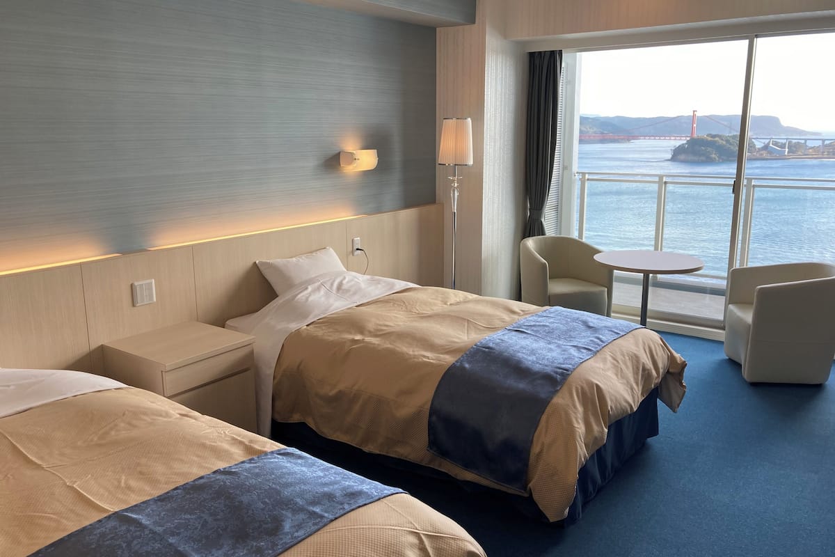 Habitación de hotel con dos camas, una mesita de noche, una lámpara, una silla, una pequeña mesa y un amplio ventanal con vistas a un puente y al agua.