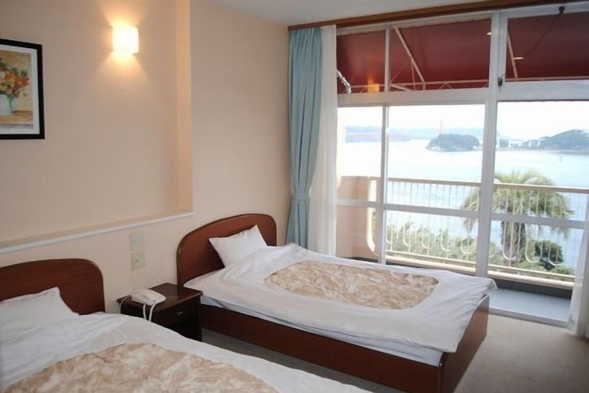 Une chambre d’hôtel avec deux lits, une grande fenêtre donnant sur la mer, un tableau au mur et une lampe de chevet.