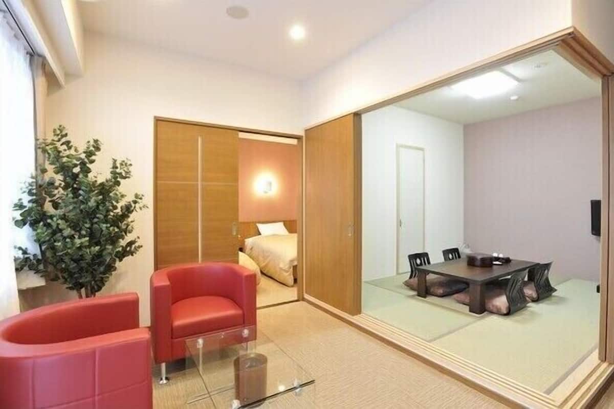Una habitación de hotel con una cama, una silla roja, una mesita de vidrio y un amplio ventanal corredero.