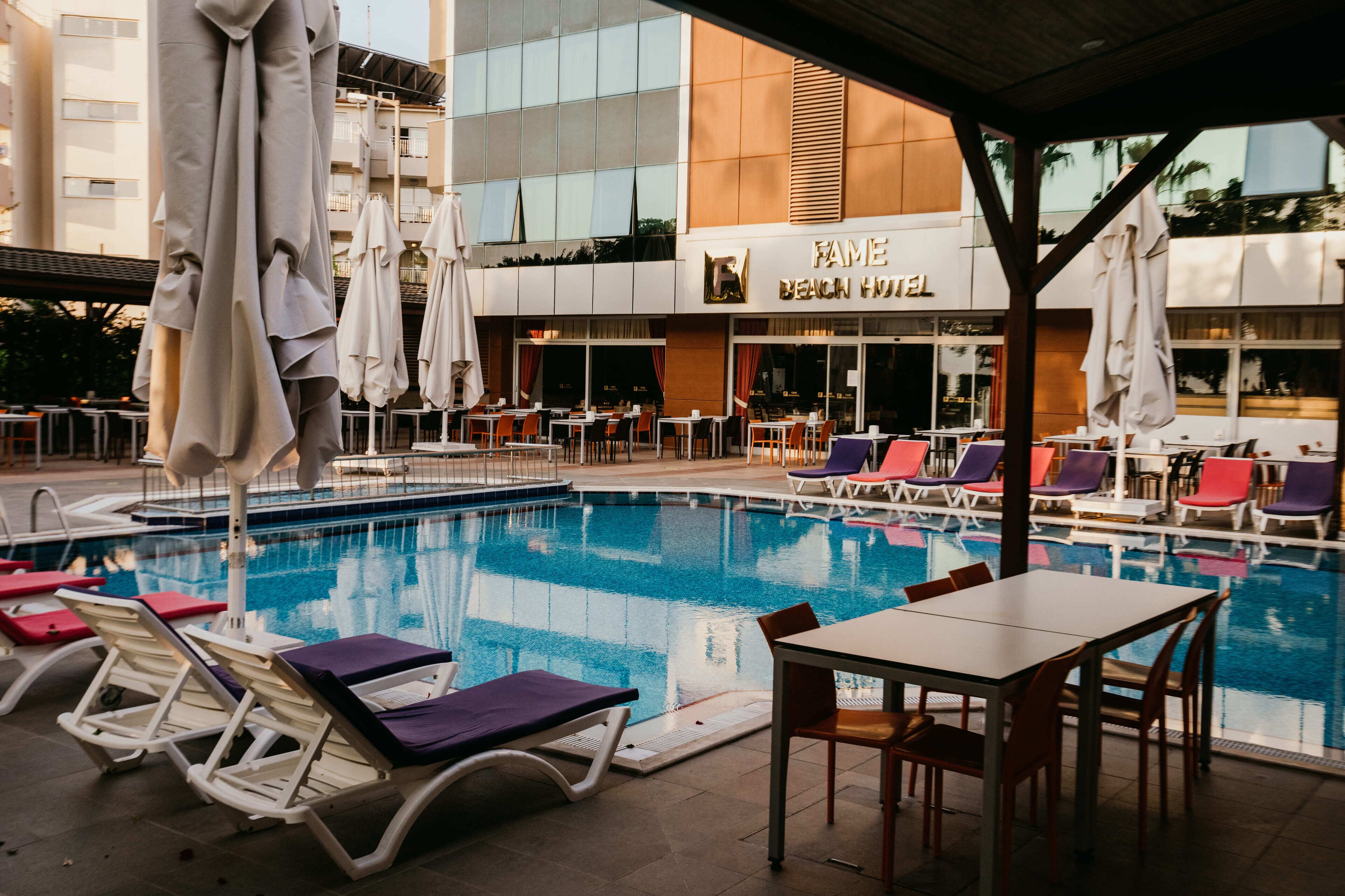 Un área de piscina con sillones de descanso y mesas, un espacio de estar techado, y un edificio con un cartel que dice "FAME BEACH HOTEL".