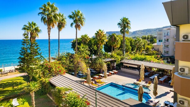 Un espace piscine aménagé sur le toit, avec des chaises longues et des parasols, offrant une vue sur un paysage côtier avec des palmiers et un ciel bleu dégagé.