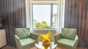Une pièce avec deux fauteuils verts, une table en verre sur laquelle se trouve un objet jaune, et une fenêtre donnant sur des palmiers et la mer.