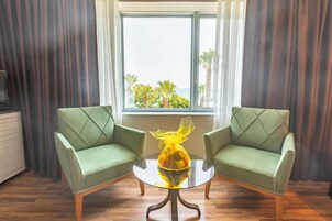 A room with two green armchairs, a glass table with a yellow object, and a window with a view of palm trees and the sea.
