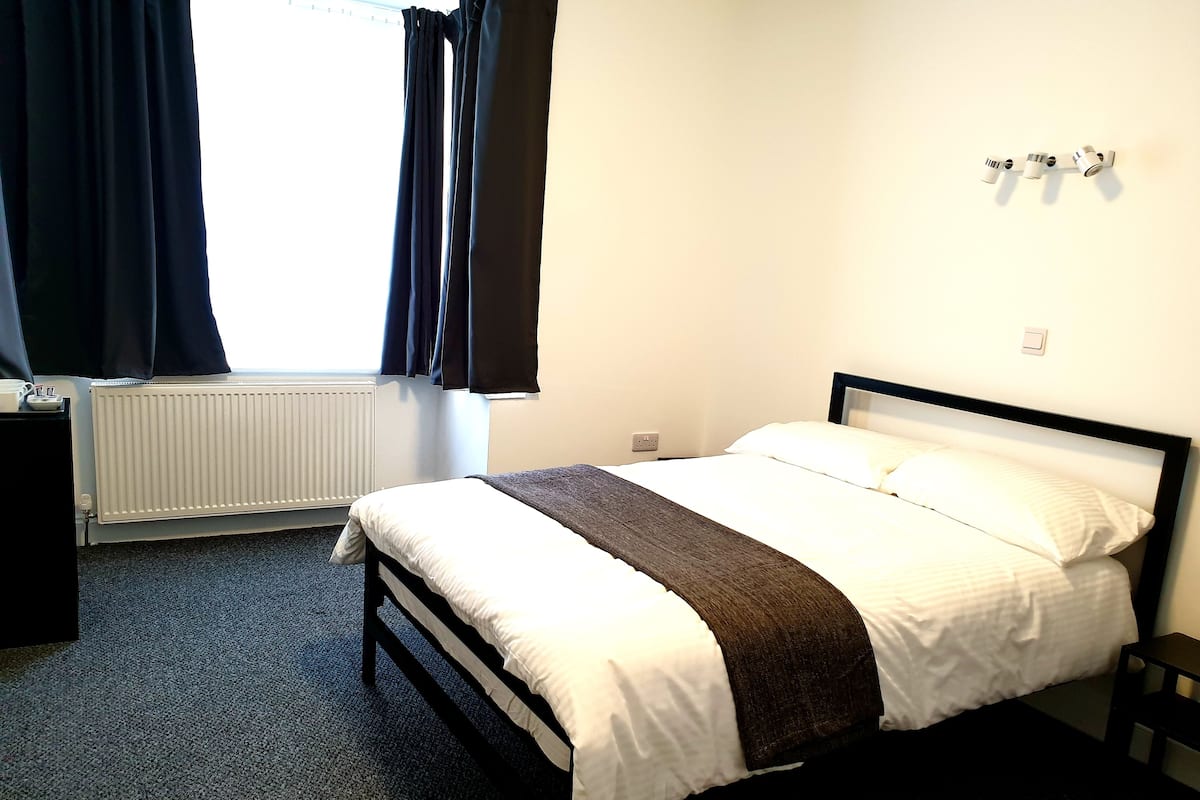 A hotel room with a bed, a bedside table, a radiator, and a window with curtains.