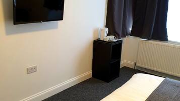 A hotel room with a bed, a flat-screen TV mounted on the wall, a bedside table with a kettle and cups, and a window with dark curtains.