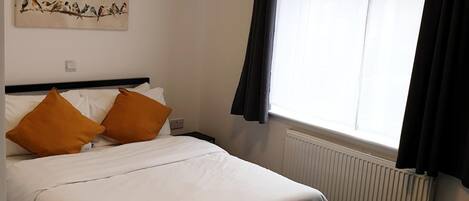 Una cama bien hecha con sábanas blancas y dos almohadas anaranjadas, un radiador, una ventana con cortinas negras y una lámpara fijada a la pared.