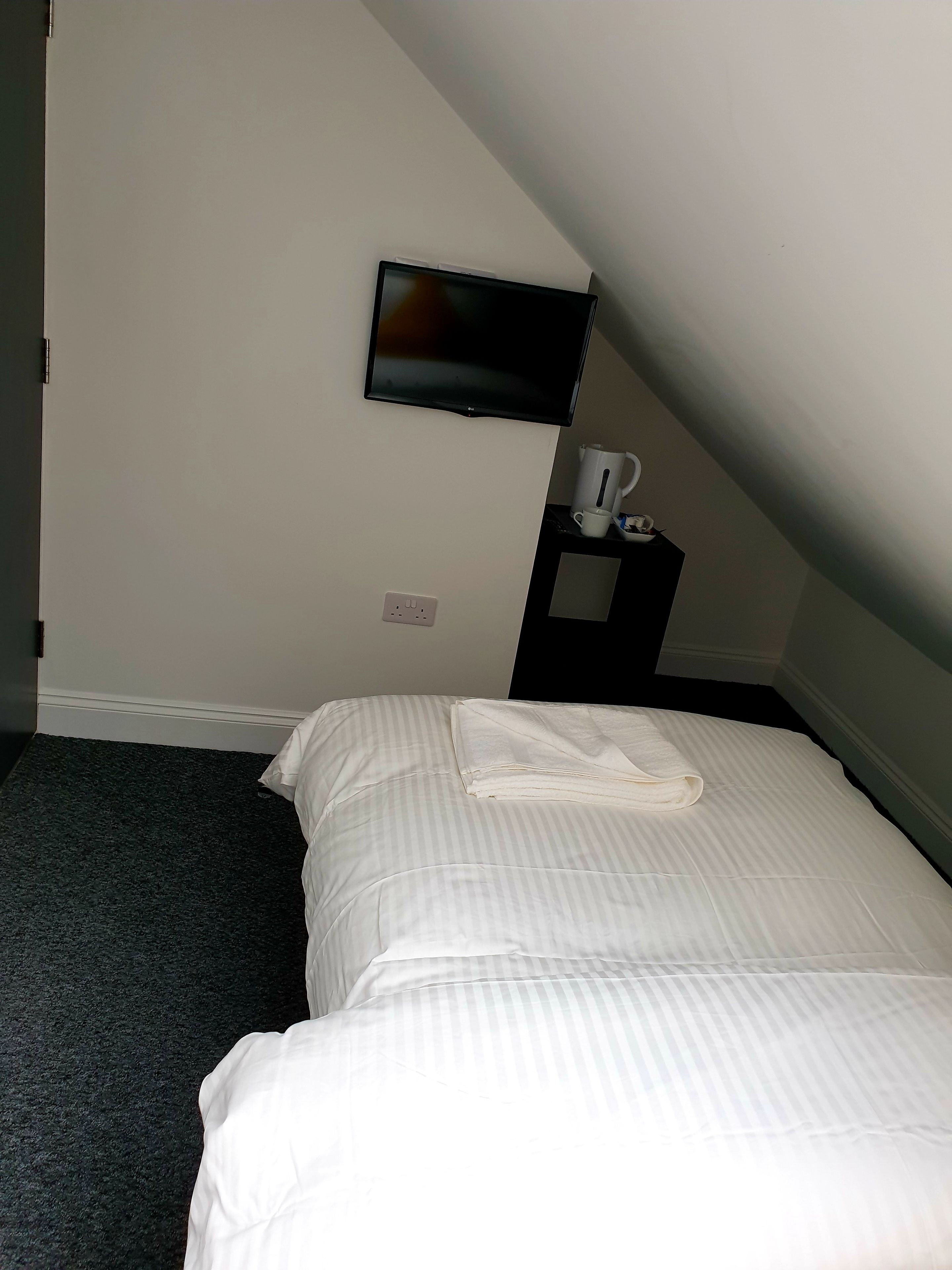 A hotel room with a bed, a TV mounted on the wall, a small table with a cup and a kettle, and a power outlet on the wall.