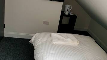 A hotel room with a bed, a TV mounted on the wall, a small table with a cup and a kettle, and a power outlet on the wall.