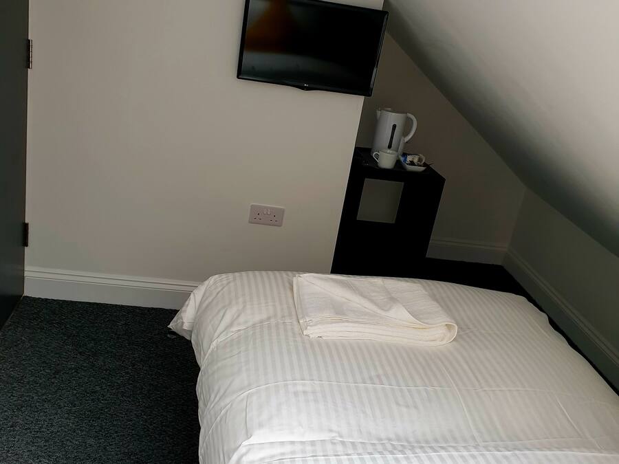 A hotel room with a bed, a TV mounted on the wall, a small table with a cup and a kettle, and a power outlet on the wall.