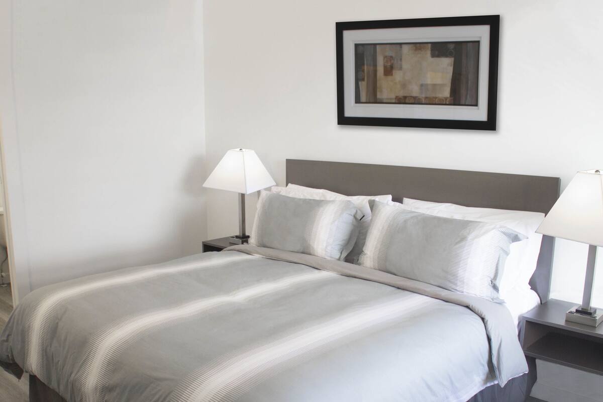 Een netjes opgemaakt bed met een grijs-witte streepdeken, twee nachtlampjes en een lijst met een abstract schilderij aan de muur.