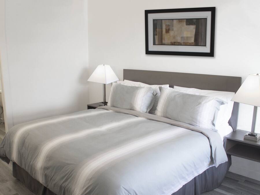 A neatly made bed with a grey and white striped bedspread, two bedside lamps, and a framed abstract painting on the wall.