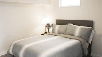 A neatly made bed with a grey bedspread and pillows, a bedside table with a lamp and a vase of flowers, and a window providing natural light.