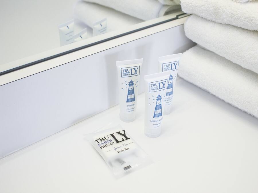 A bathroom with a mirror reflecting a stack of white towels and three bottles of TruLY body wash and conditioner.