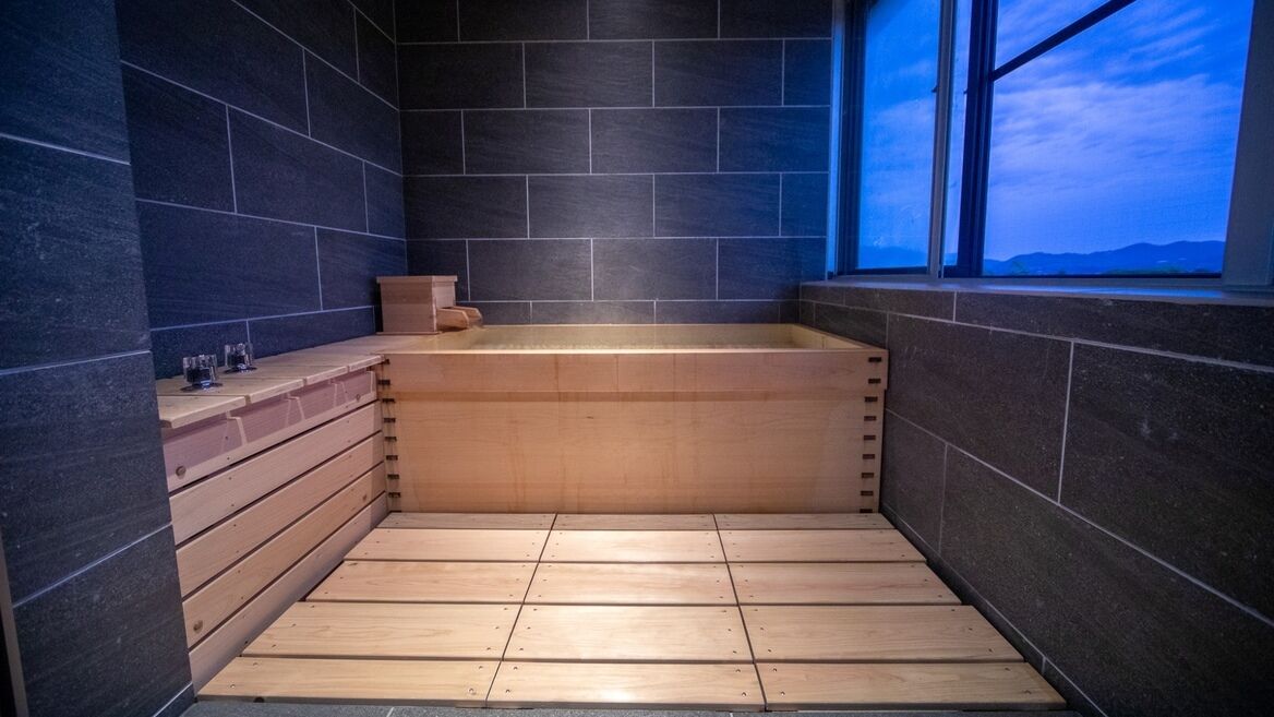 Ein modernes Badezimmer mit einer Holzwanne, dunklen Fliesenwänden und einem Fenster mit Ausblick.