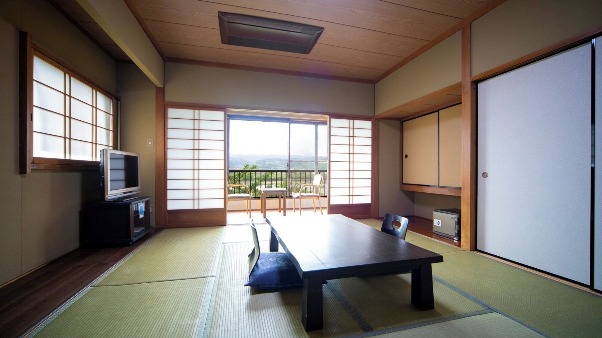 Ein traditioneller japanischer Raum mit Tatami-Böden, einem niedrigen Tisch, Stühlen, einem Fernseher und Blick ins Freie.