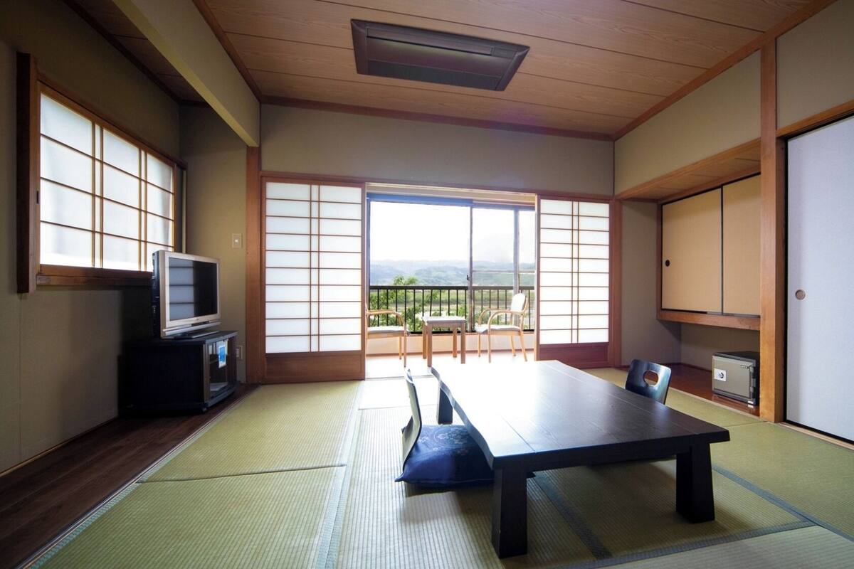 Um quarto japonês tradicional com tatames, uma mesa baixa, cadeiras, televisão e vista para o exterior.