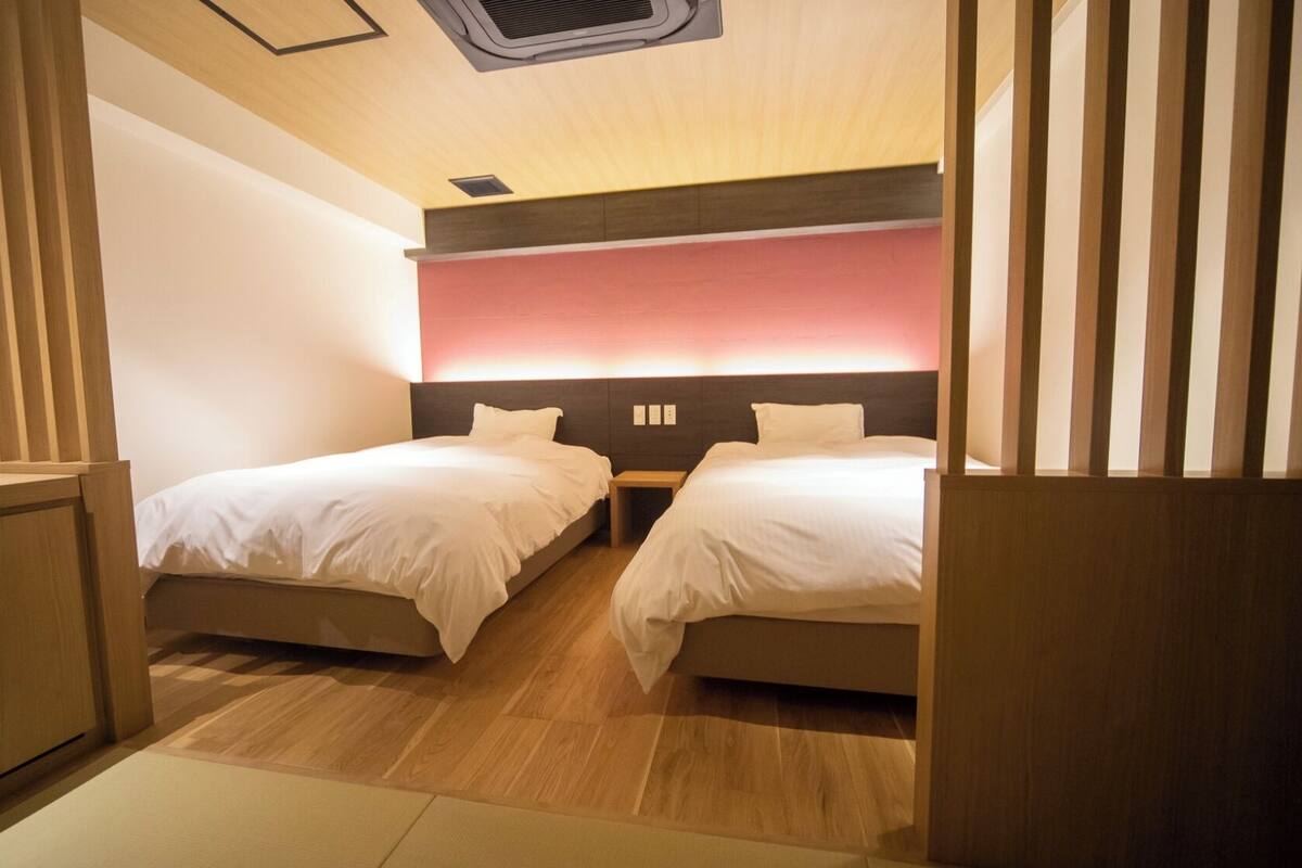 Quarto de hotel com duas camas, piso de madeira, um respiradouro no teto e um candeeiro fixado na parede.
