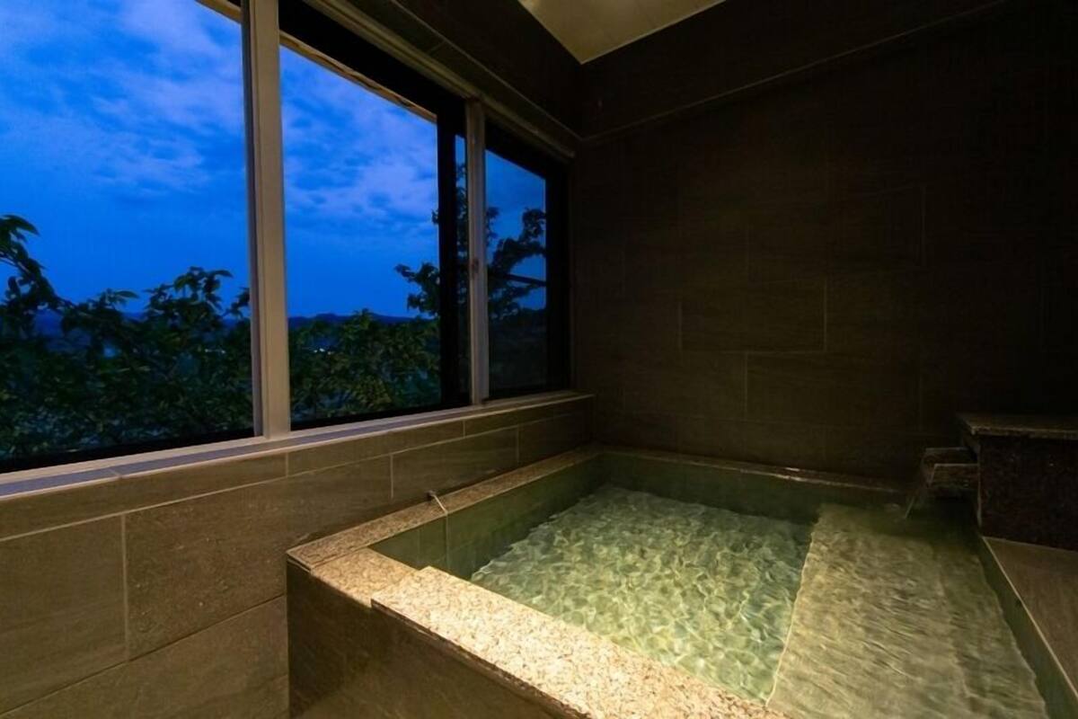 Um salão de spa com uma janela ampla, uma banheira de hidromassagem e vista para árvores.