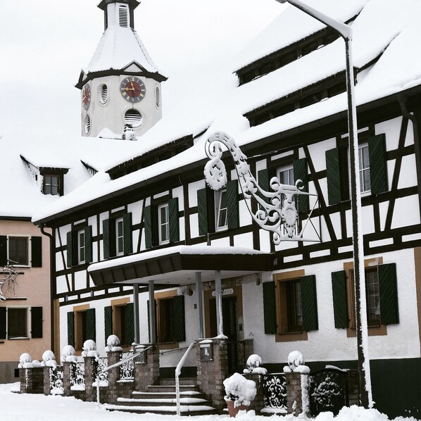 Un edificio tradicional con entramado de madera, fachada cubierta de nieve y una torre del reloj al fondo.