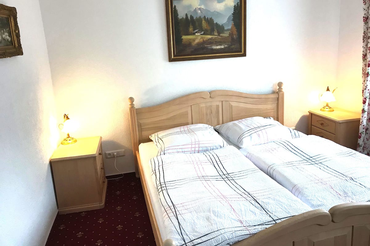 A double bed with a wooden headboard, two bedside tables with lamps, a painting on the wall, and a floral curtain.