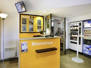 A hotel reception area with a yellow counter, a reception desk, a television mounted on the wall, a vending machine, and a small plant. - hotelF1 Brive Ussac (Ussac)