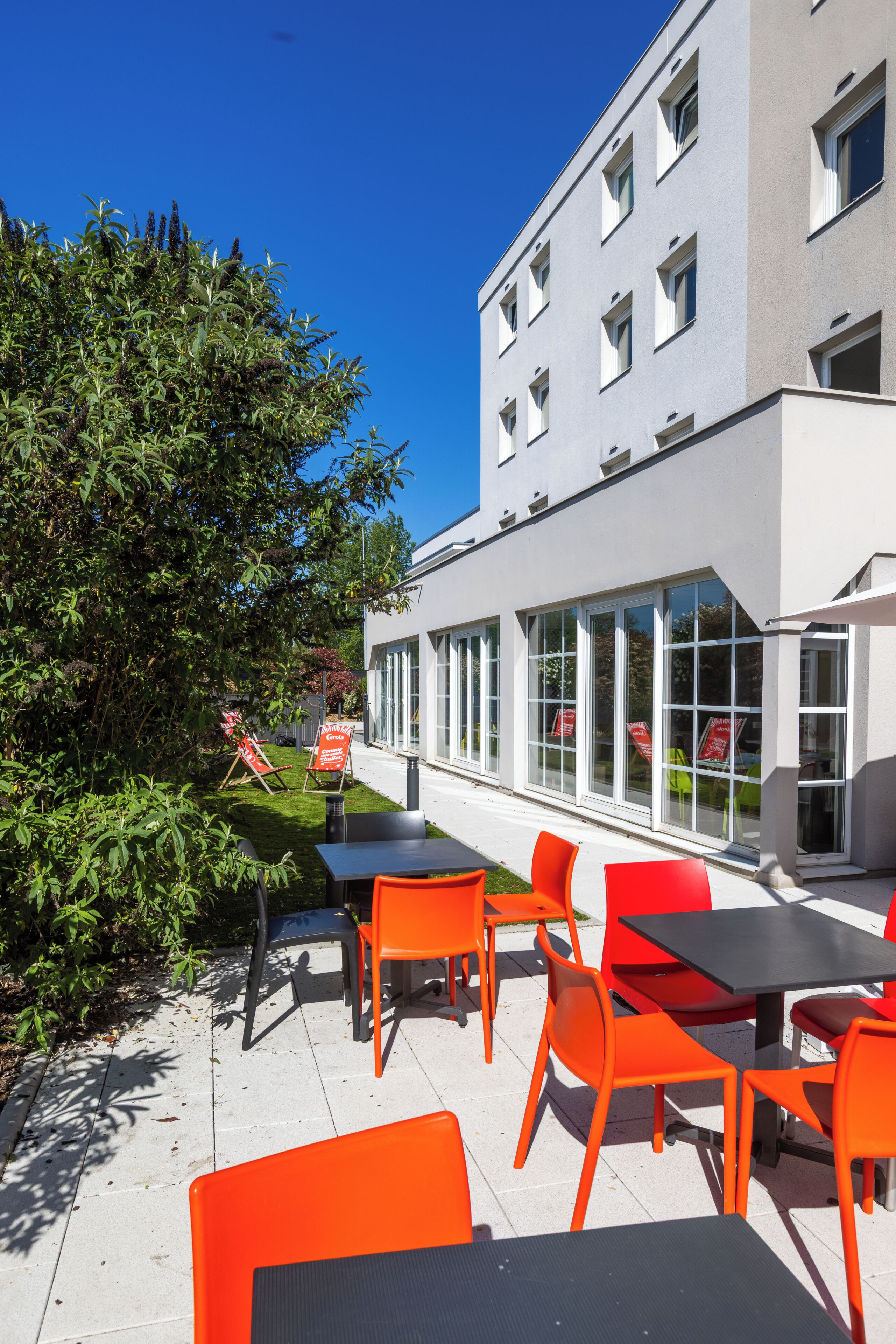 An outdoor seating area with orange chairs and tables, a building with large windows, and a clear blue sky.