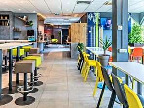 A modern restaurant with high tables and chairs, a bar area, and a seating section with colorful chairs. - ibis budget Strasbourg Sud Illkirch Geispolsheim (Geispolsheim)