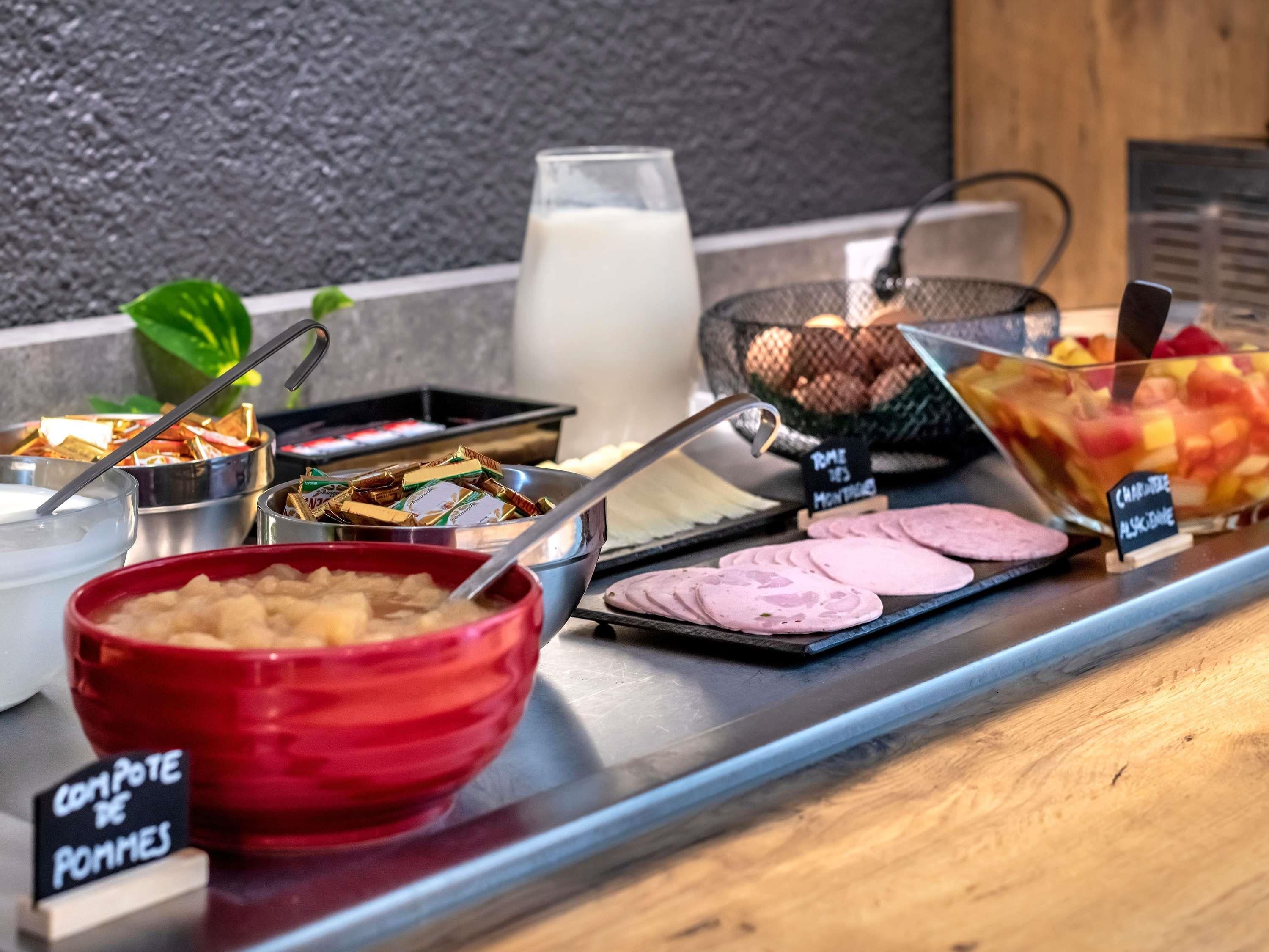 Een ontbijtbuffet met diverse etenswaren, waaronder een rode schaal met 'COMPOTE DE POMMES', een glas melk en een schaal fruit.