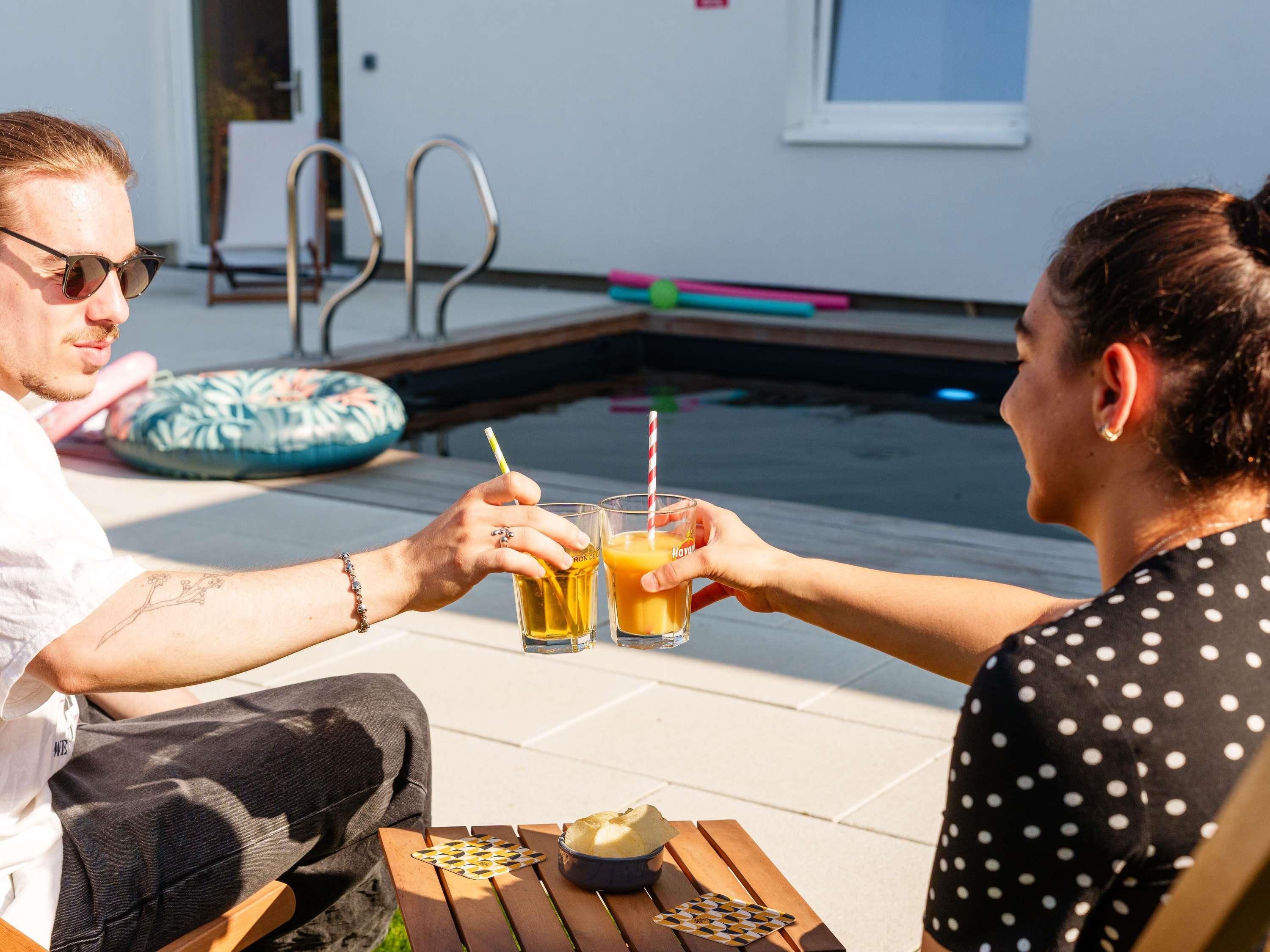 Dos personas brindan con bebidas en una mesa junto a la piscina, con sillas de descanso y la piscina al fondo.