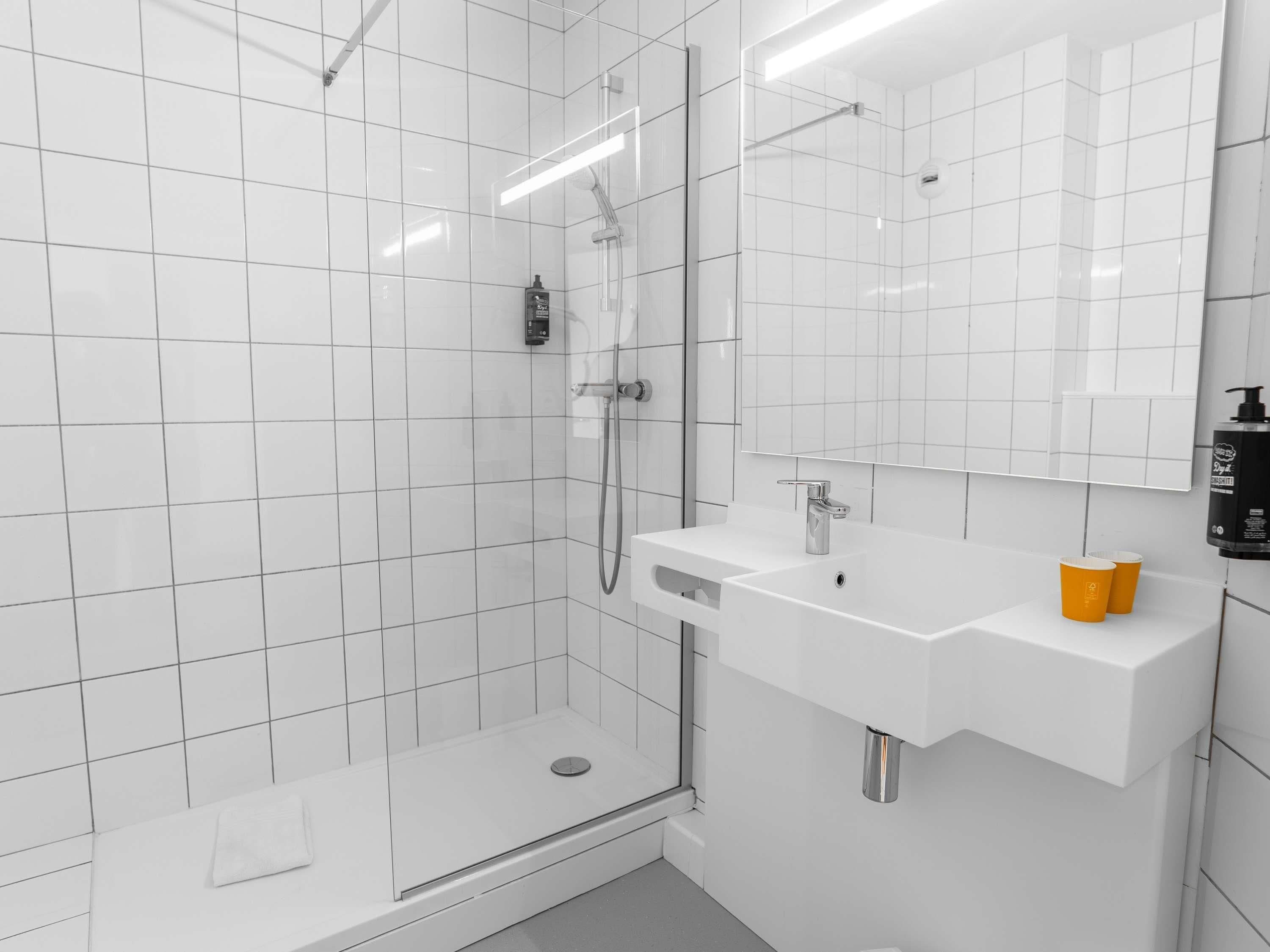 Um banheiro moderno com azulejos brancos, box de vidro, pia branca com espelho e uma xícara amarela no balcão.