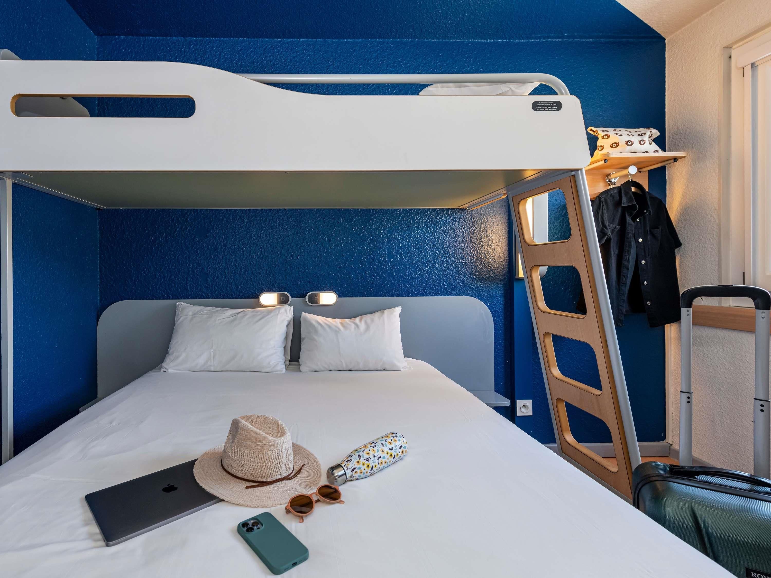 Habitación con literas, una escalera, una maleta, un sombrero, gafas de sol y un ordenador portátil sobre la cama.