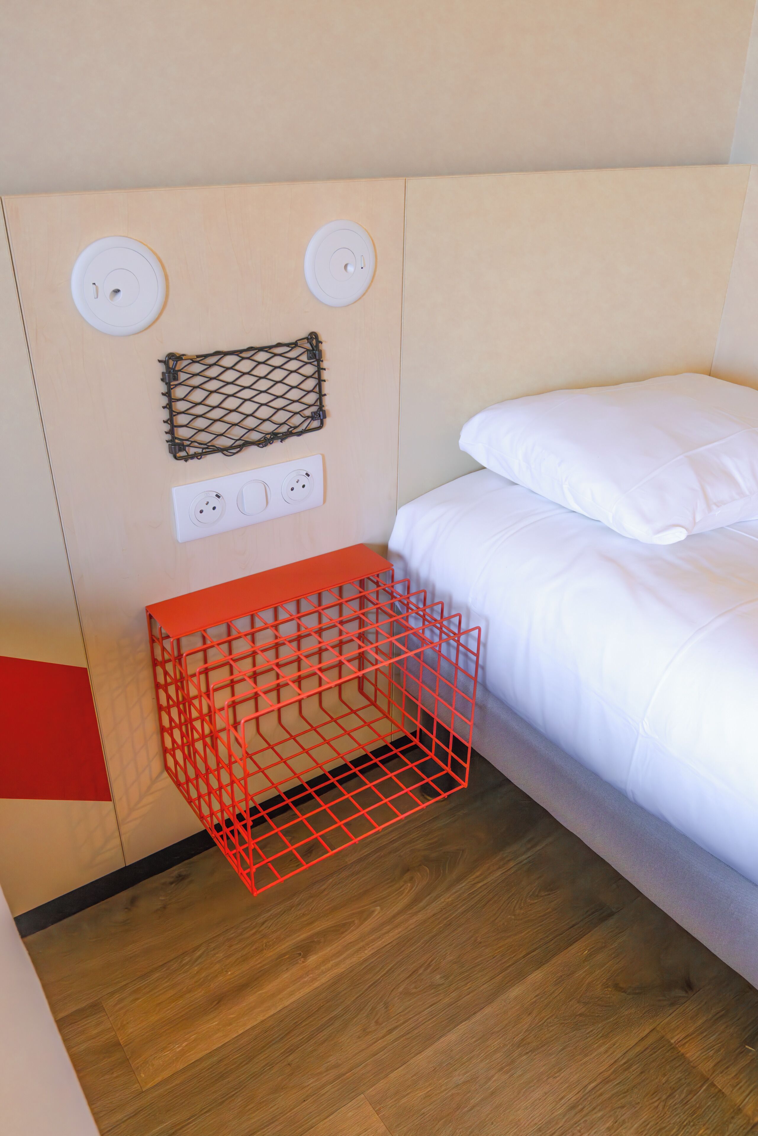 A bedroom with a bed, a red storage bin, a power outlet, and a wall-mounted shelf.