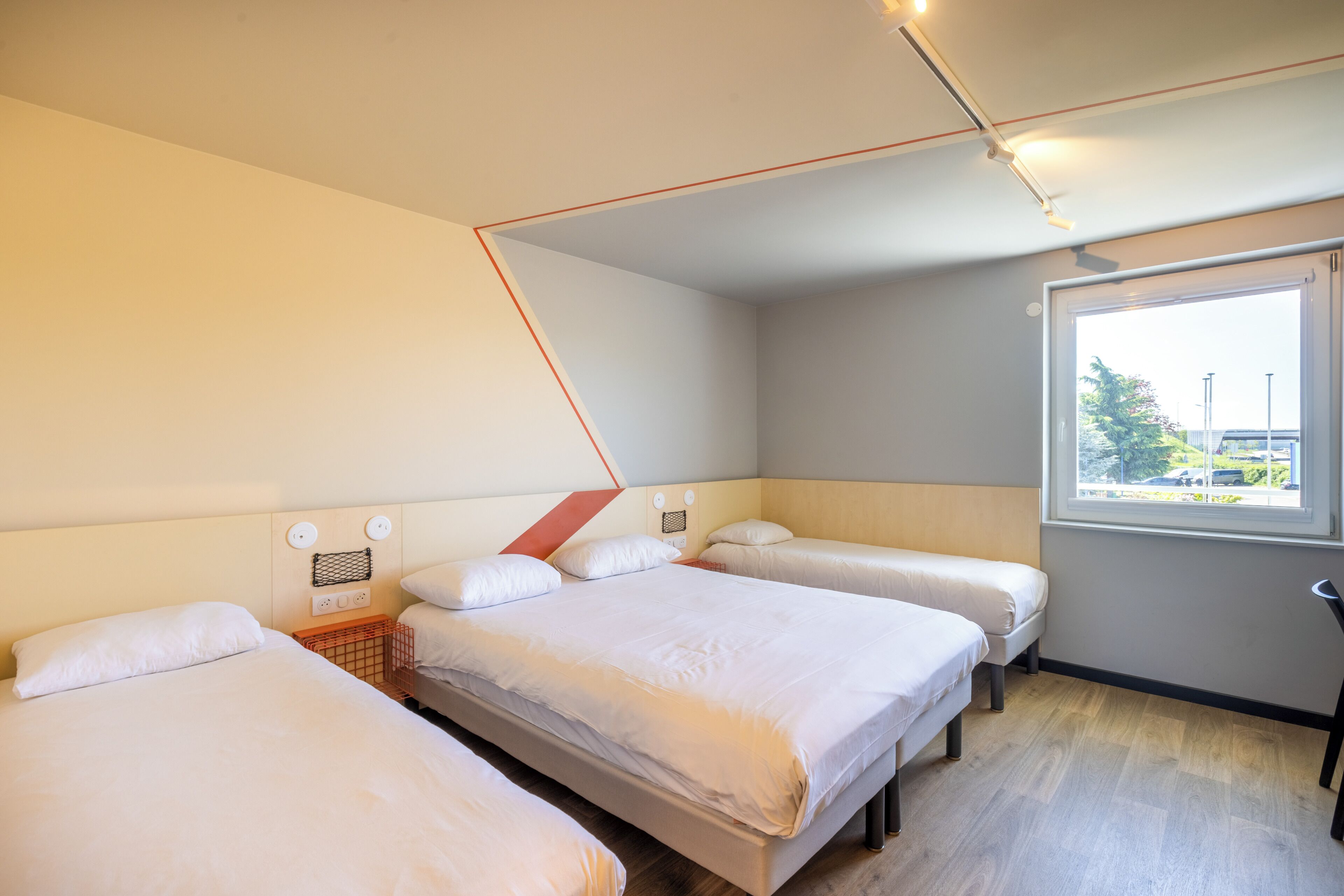 A hotel room with three beds, a window with a view, and a ceiling with exposed ductwork.
