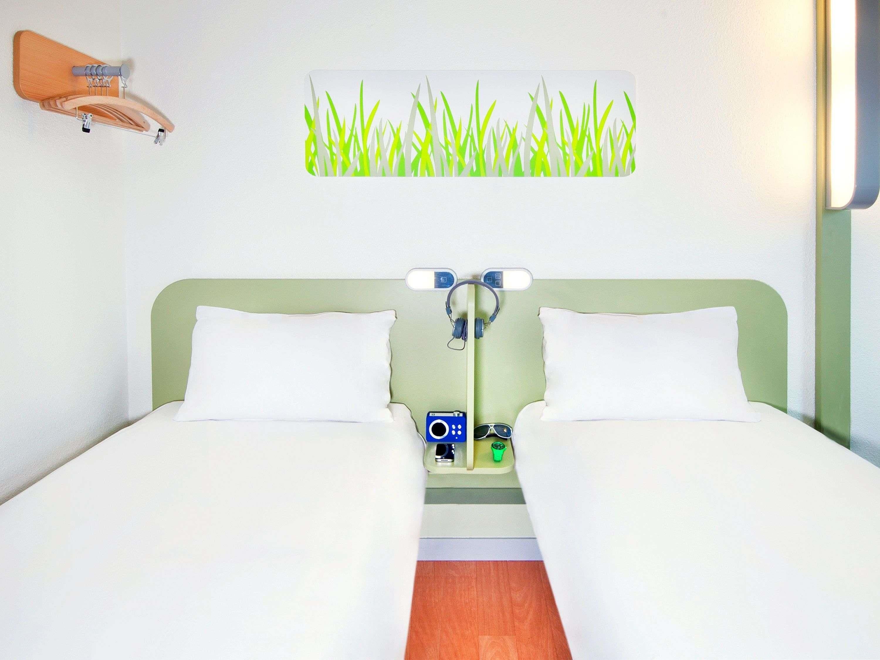 A double bed with white linens, a wall-mounted clock, and a small table with a remote control and sunglasses.