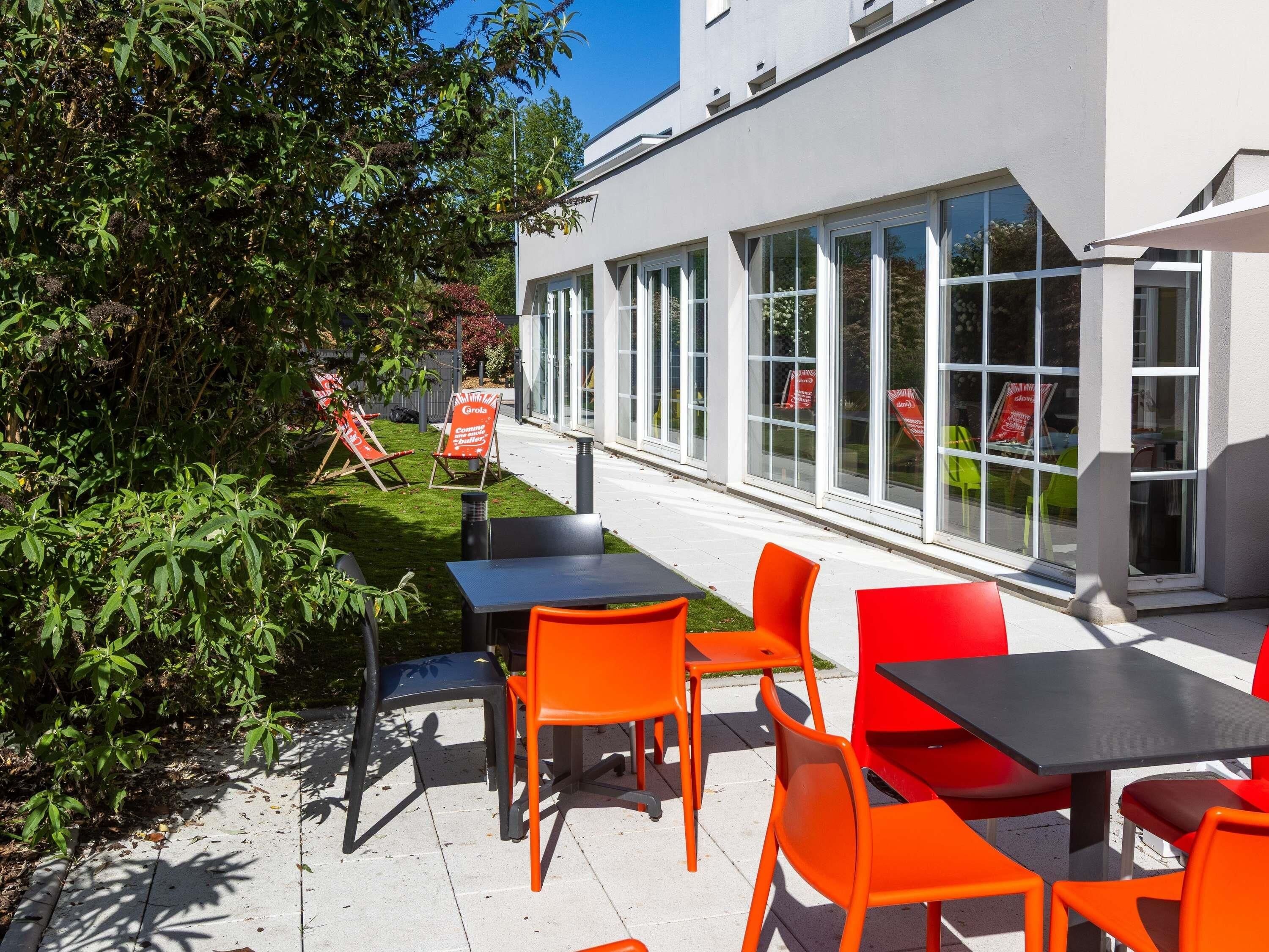 Un espace extérieur aménagé avec des tables et des chaises, un bâtiment doté de grandes fenêtres et une zone verdoyante avec des plantes.