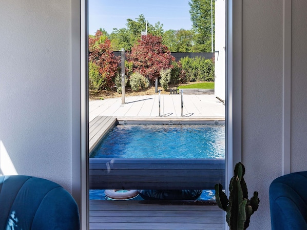 Une piscine entourée d’une terrasse en bois, un cactus et une vue sur des arbres et une clôture.