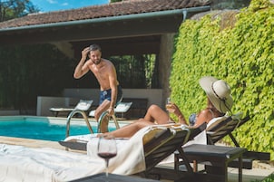 Zona junto a la piscina con sillones de descanso, un área de estar techada y un seto verde exuberante.