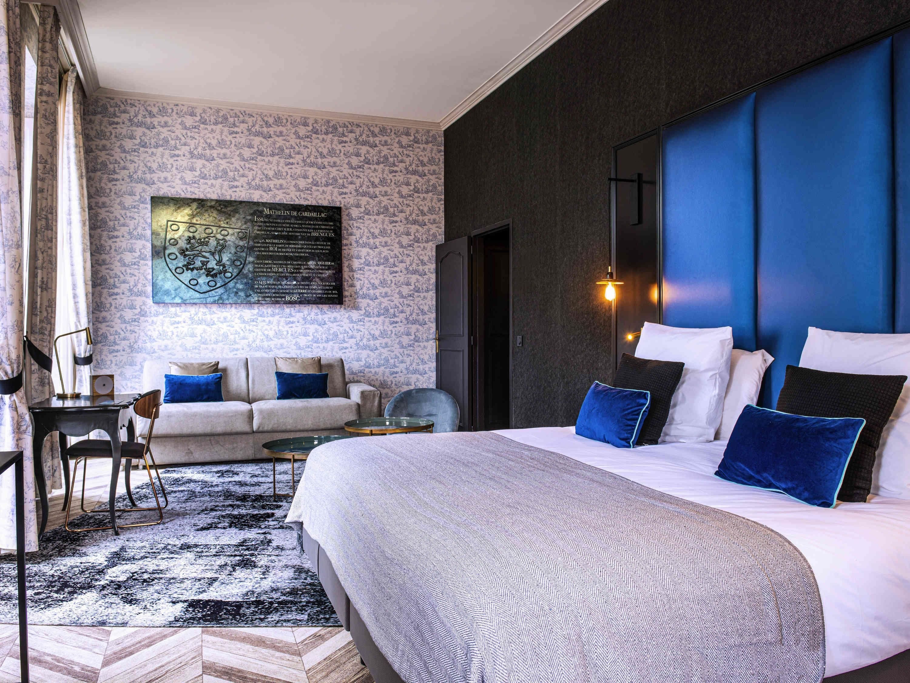 A modern hotel room with a large bed, a seating area with a sofa and armchairs, a desk, and a wall-mounted artwork.