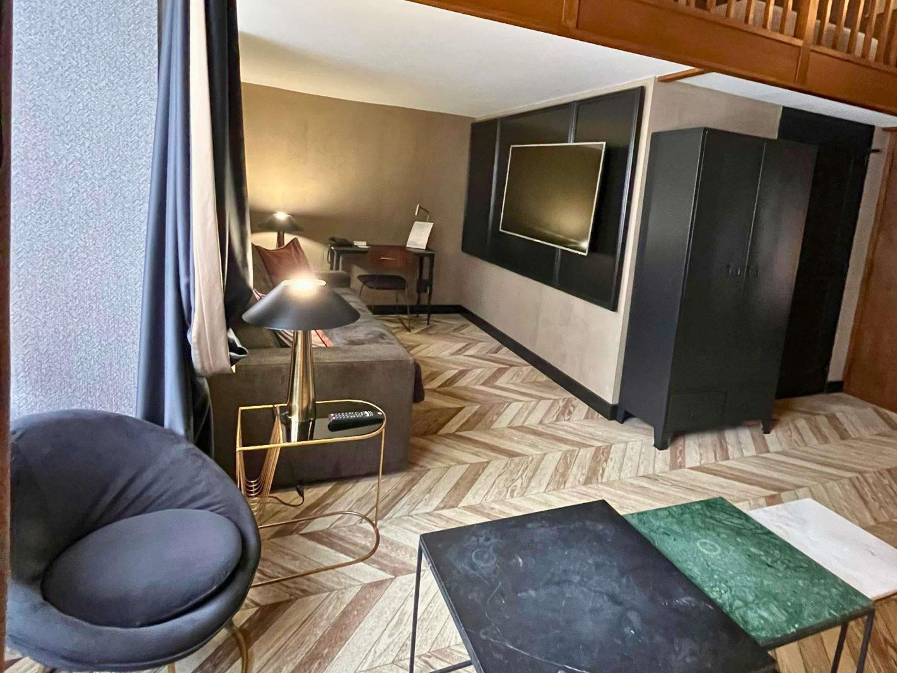 A modern hotel room with a sofa, a chair, a small table, a TV mounted on the wall, and a wardrobe.