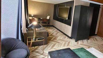 A modern hotel room with a sofa, a chair, a small table, a TV mounted on the wall, and a wardrobe.